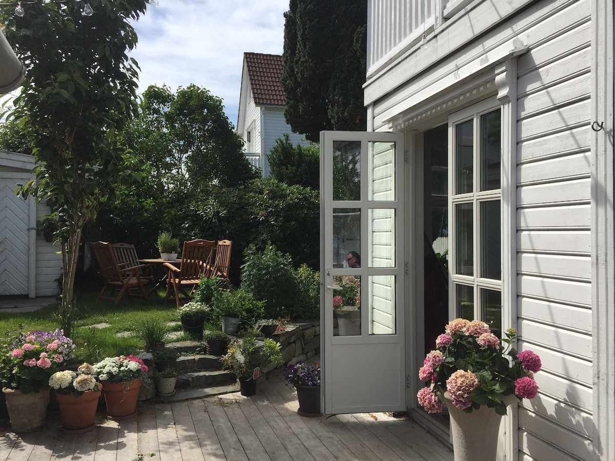 A white wooden door stands open, leading to a garden area filled with various potted plants and flowers. Two wooden chairs are positioned among the greenery, creating a cozy outdoor seating area. Sunlight filters through the trees, adding a warm ambiance.