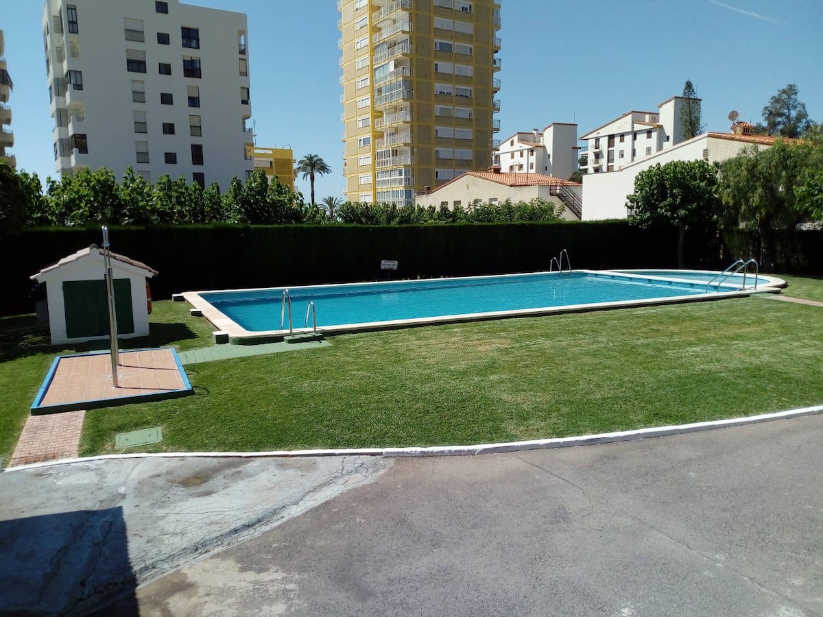 A spacious outdoor area is presented, featuring a large swimming pool surrounded by well-maintained grass. Additional amenities include a small building nearby and lounge chairs positioned along the poolside. Tall residential buildings frame the scene, indicating a community setting.