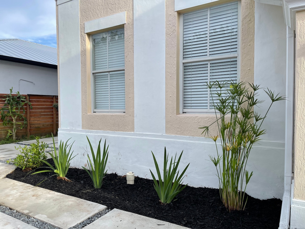 A well-maintained exterior features two large windows with white shutters, surrounded by vibrant greenery. The landscape includes a mix of tall plants and low shrubs, complemented by black mulch, providing a neat and fresh appearance.