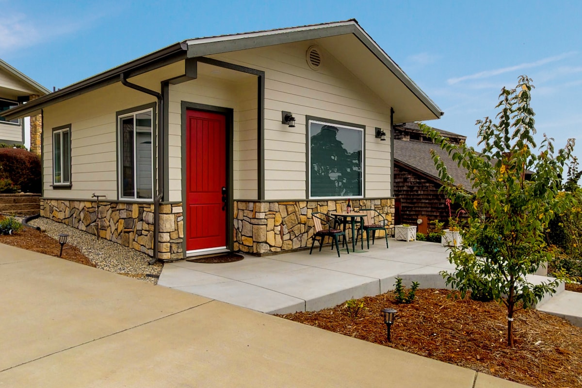 A modern home exterior is highlighted by a vibrant red door and stone accents on the facade. A small patio area with two chairs and a table is positioned under a large window, while landscaped areas with plants enhance the welcoming entrance.
