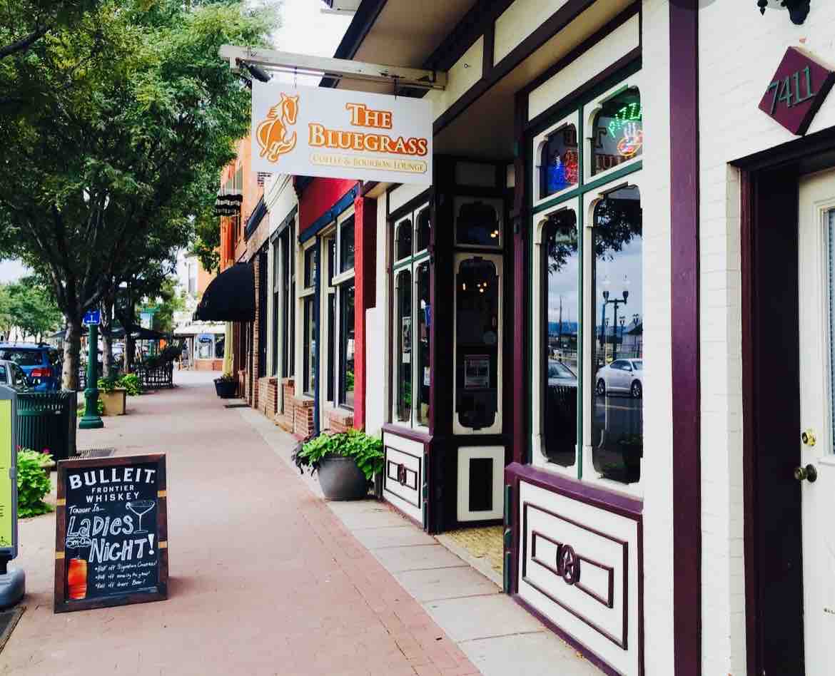 A vibrant streetscape showcases a charming exterior of a restaurant with large windows and decorative signage. Planters with greenery line the sidewalk, contributing to a lively atmosphere. Nearby storefronts and awnings enhance the inviting ambiance of this bustling neighborhood.