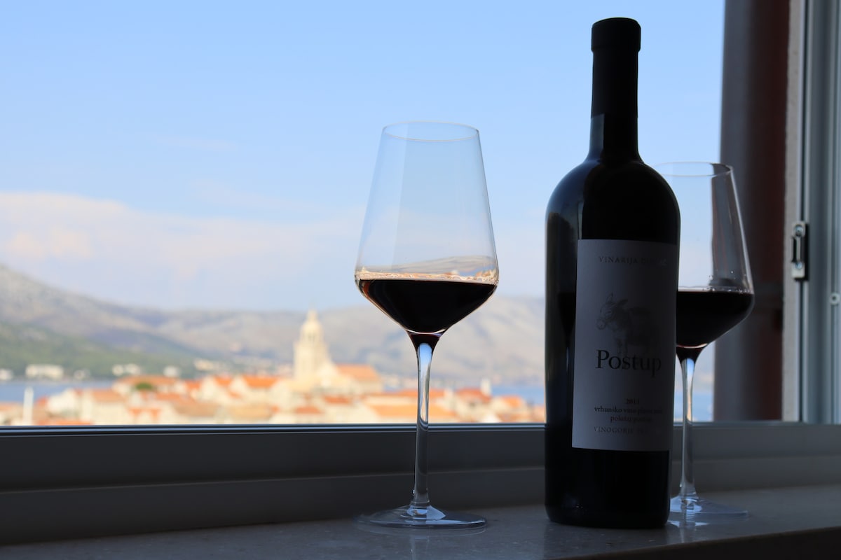 A bottle of red wine is displayed with two glasses filled to highlight a scenic view of the city, featuring historic buildings and the distant landscape. The image captures the essence of relaxation and enjoyment, framed by a window that invites the outside scenery in.