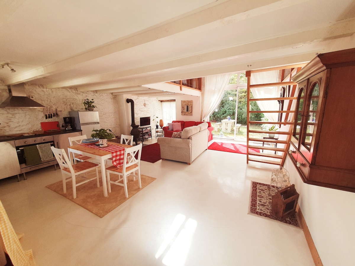 An open-plan living area showcases a well-equipped kitchen, a dining table with four chairs, and a comfortable sofa facing a wood-burning stove. Large windows allow natural light to flood the space, with soft drapes enhancing the serene atmosphere. A staircase leads to an upper level.