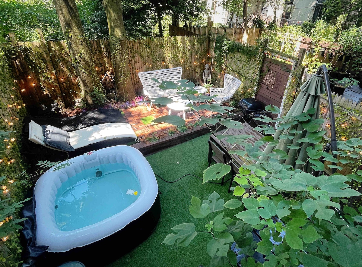 A serene backyard garden is surrounded by lush greenery and privacy fencing. An inflatable hot tub sits to the side, while comfortable seating areas, adorned with twinkling lights, invite relaxation. A small wooden deck offers additional space for lounging in this tranquil outdoor setting.