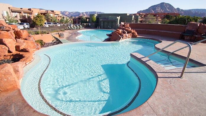 A large, inviting pool meanders through a landscaped area, framed by natural rock features. Comfortable lounge chairs are positioned along the water's edge. In the distance, mountains provide a scenic backdrop to the serene setting.