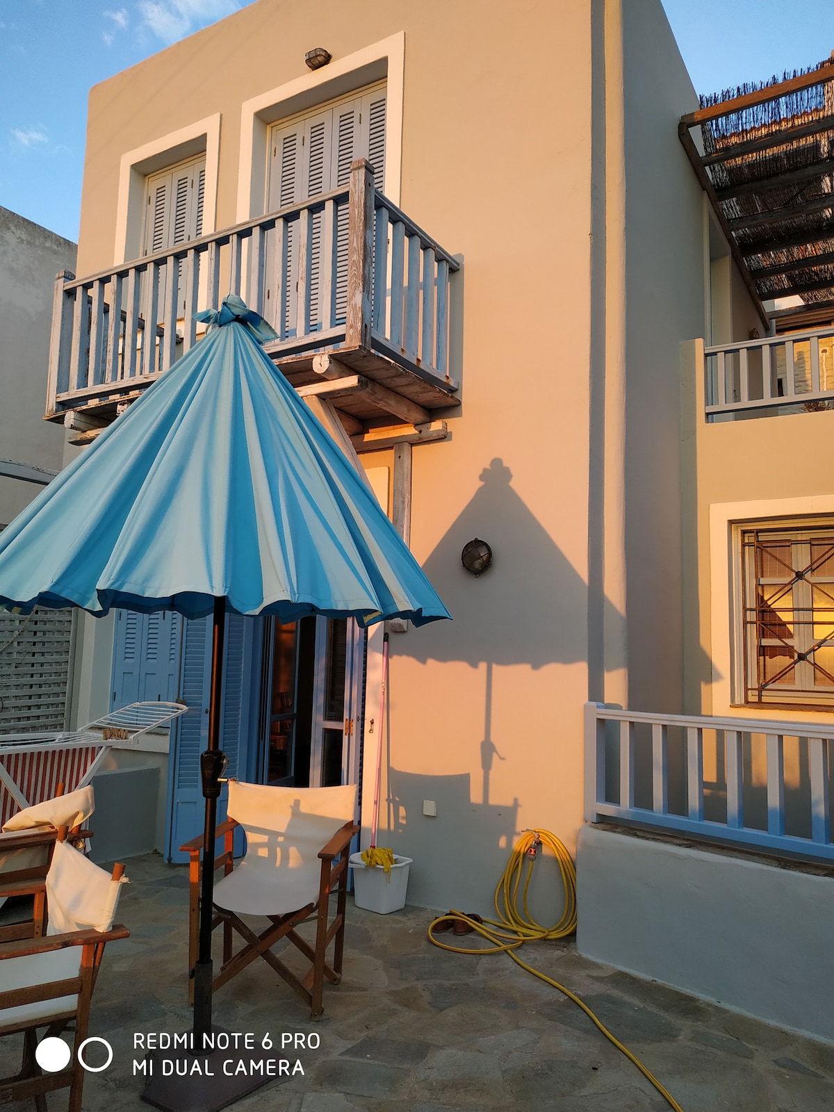 The exterior of a three-level home is shown, featuring light-colored walls and blue window shutters. A spacious balcony is supported by wooden posts, while a large blue umbrella provides shade over outdoor seating. The ground is paved with stone, creating a welcoming entrance.