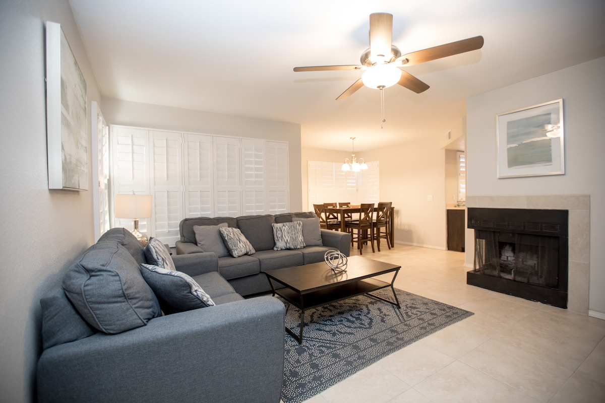 The living area features a spacious sectional sofa with decorative pillows, accompanied by a sleek coffee table on a patterned area rug. Light enters through the windows covered with wooden shutters, and a ceiling fan is present above, enhancing airflow in the room.