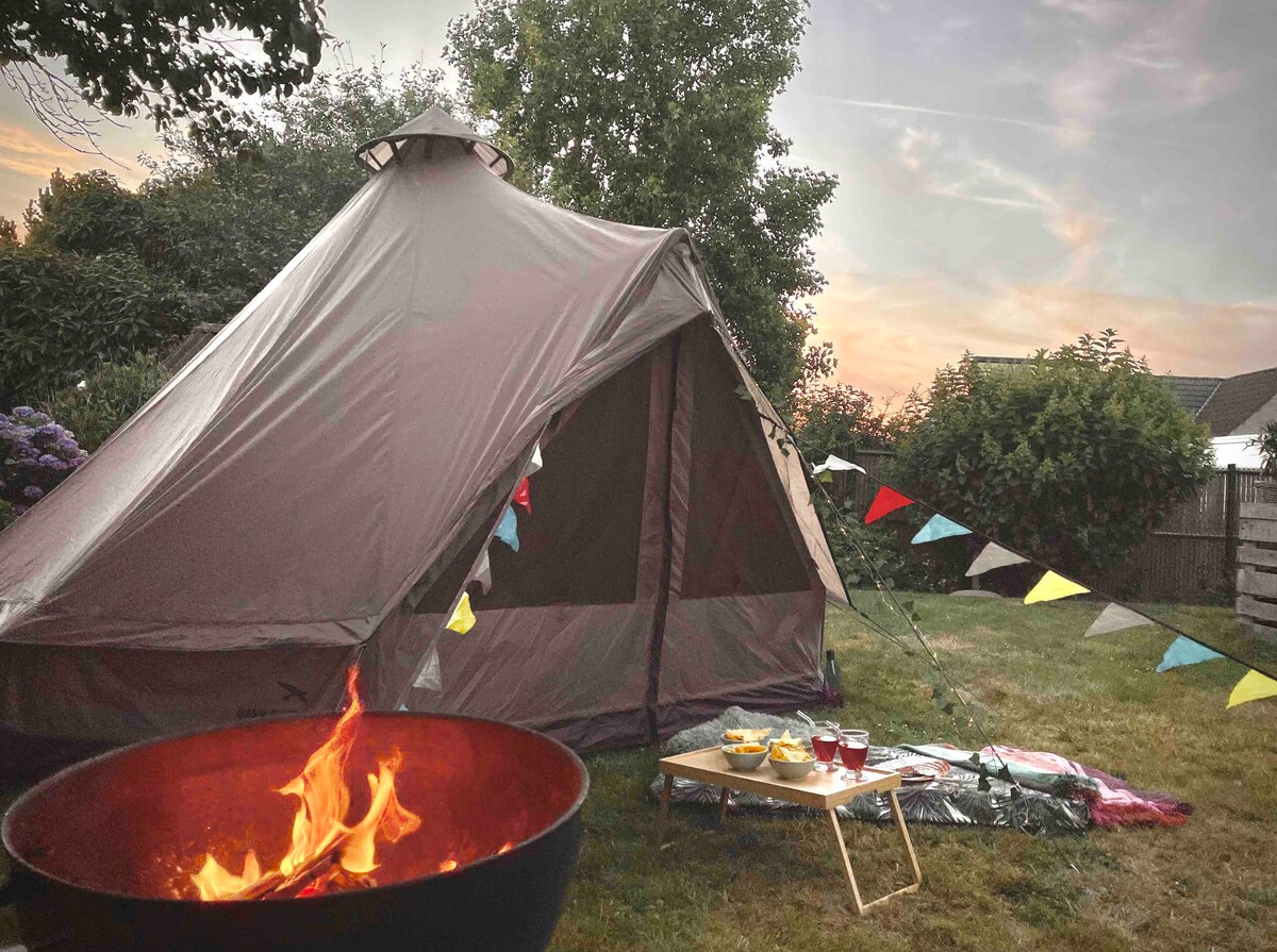A glamping tent is set within a garden, accompanied by a circular fire pit with flames visible. Colorful bunting is strung nearby, and a picnic setup with a low table, snacks, and drinks can be seen on the ground, adding to the outdoor experience.