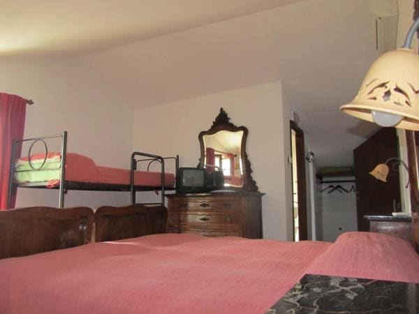 A spacious bedroom features a large bed covered with a red blanket, complemented by wooden furniture, including a dresser and a mirror. A bunk bed with green bedding is positioned against the wall, and natural light softly enters through the window dressed with red curtains.
