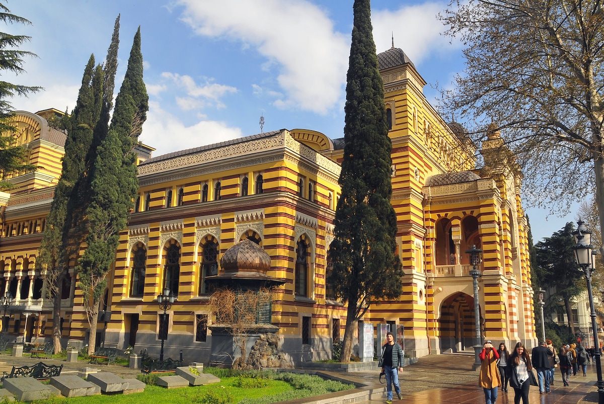 An architectural marvel stands with vibrant striped yellow and brick facades. Tall, evergreen cypress trees frame the structure, while the surrounding area features well-maintained pathways and green grass. The spacious courtyard is animated by people leisurely walking, enjoying the scenic environment.
