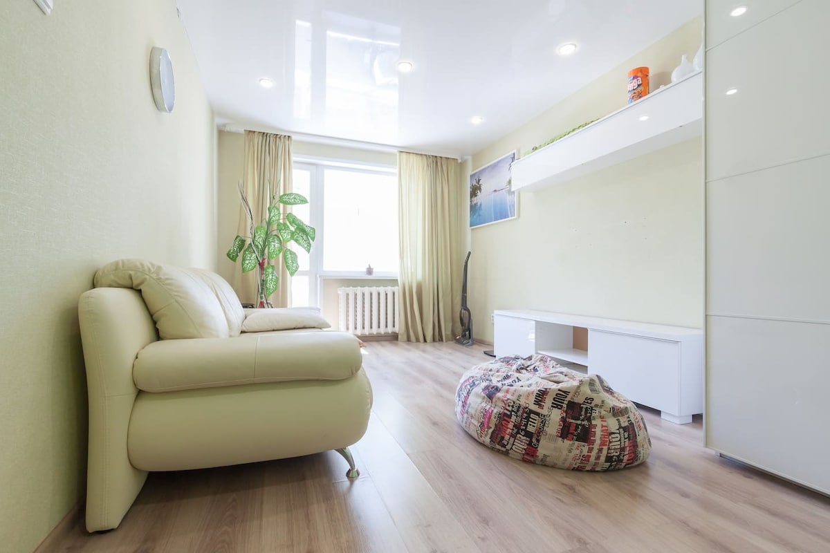 A bright living room features a light-colored leather sofa and a large window dressed with soft curtains. A plush bean bag chair is placed on the wooden floor, with a low white media console positioned against the wall. A small plant adds a touch of greenery.