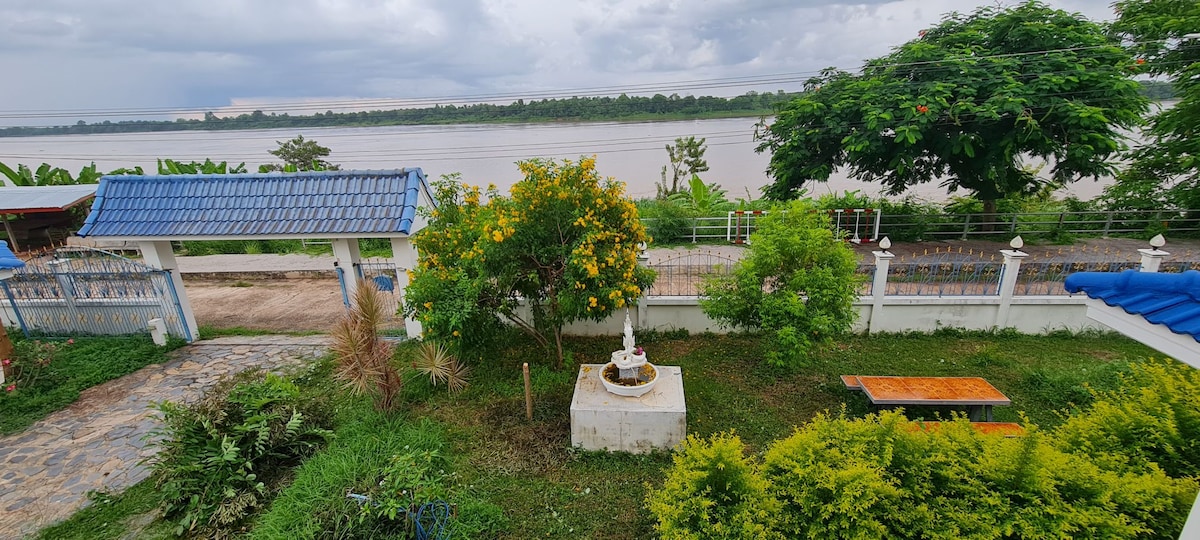 A serene outdoor area features a fountain surrounded by greenery, with a picnic table nearby. The expansive view of the river is framed by lush trees and a distant shoreline, while a pathway leads up to a gated entrance with blue roofing.