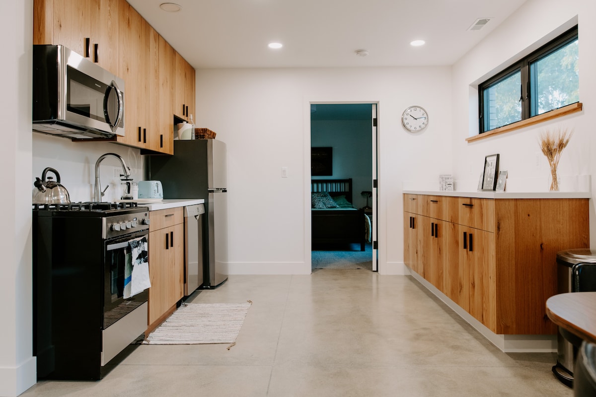 A modern kitchen features wood cabinetry and sleek stainless steel appliances, including a gas stove and refrigerator. Natural light enters through a large window, illuminating the space. The adjoining area leads to a bedroom, hinting at comfortable sleeping accommodations.