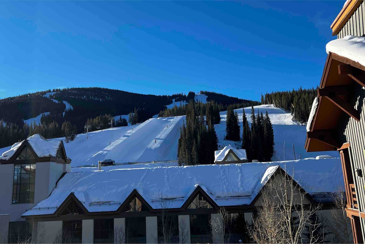 A snow-covered landscape showcases the ski runs of Copper Mountain, framed by tall evergreen trees. The clear blue sky enhances the view, while the rooftops of adjacent buildings are partially visible in the foreground.