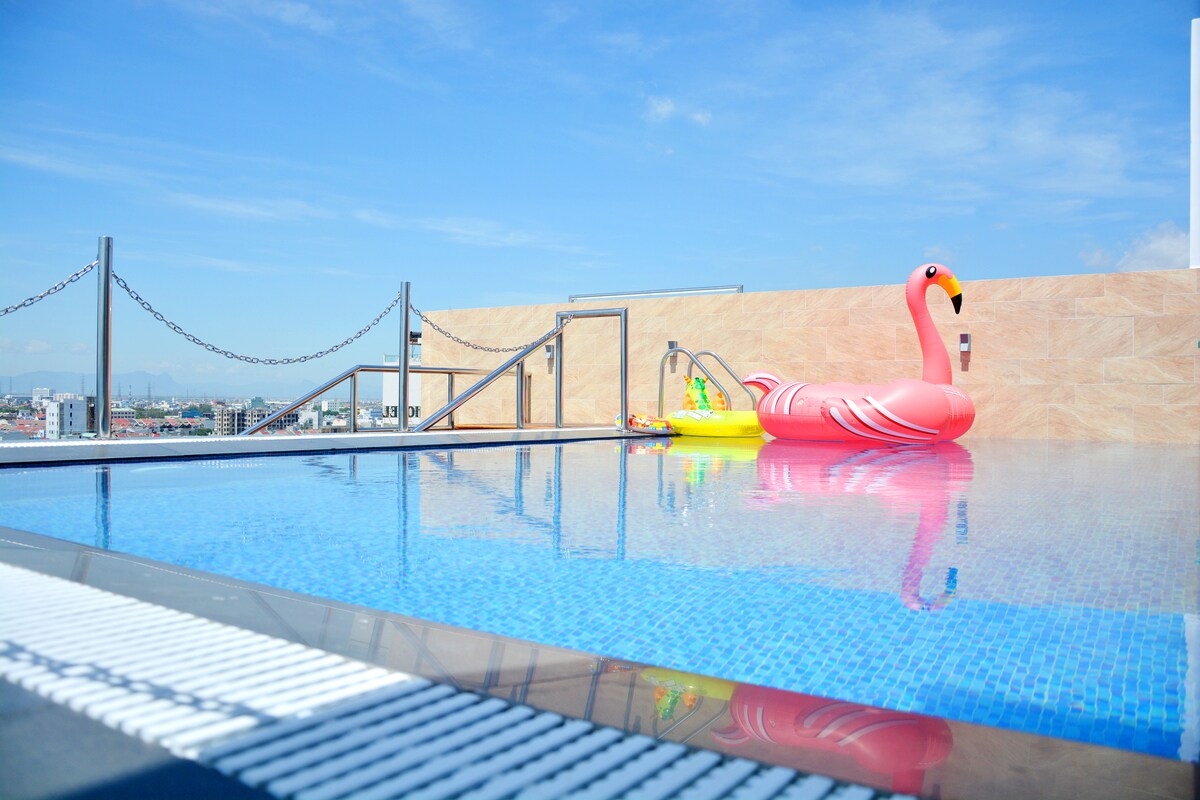 A rooftop pool features clear blue water, reflecting the bright sky. A large pink flamingo float rests on the surface, while a few colorful inflatables sit nearby. The surrounding area showcases a panoramic view of the city and distant mountains.