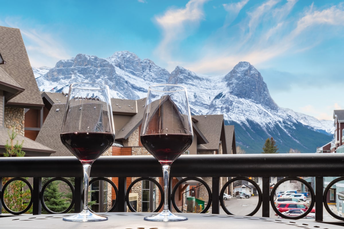 Two glasses of red wine are positioned on a balcony railing, overlooking majestic mountain peaks. The backdrop features a dramatic sky with soft clouds, and the adjacent buildings create a cozy alpine atmosphere.