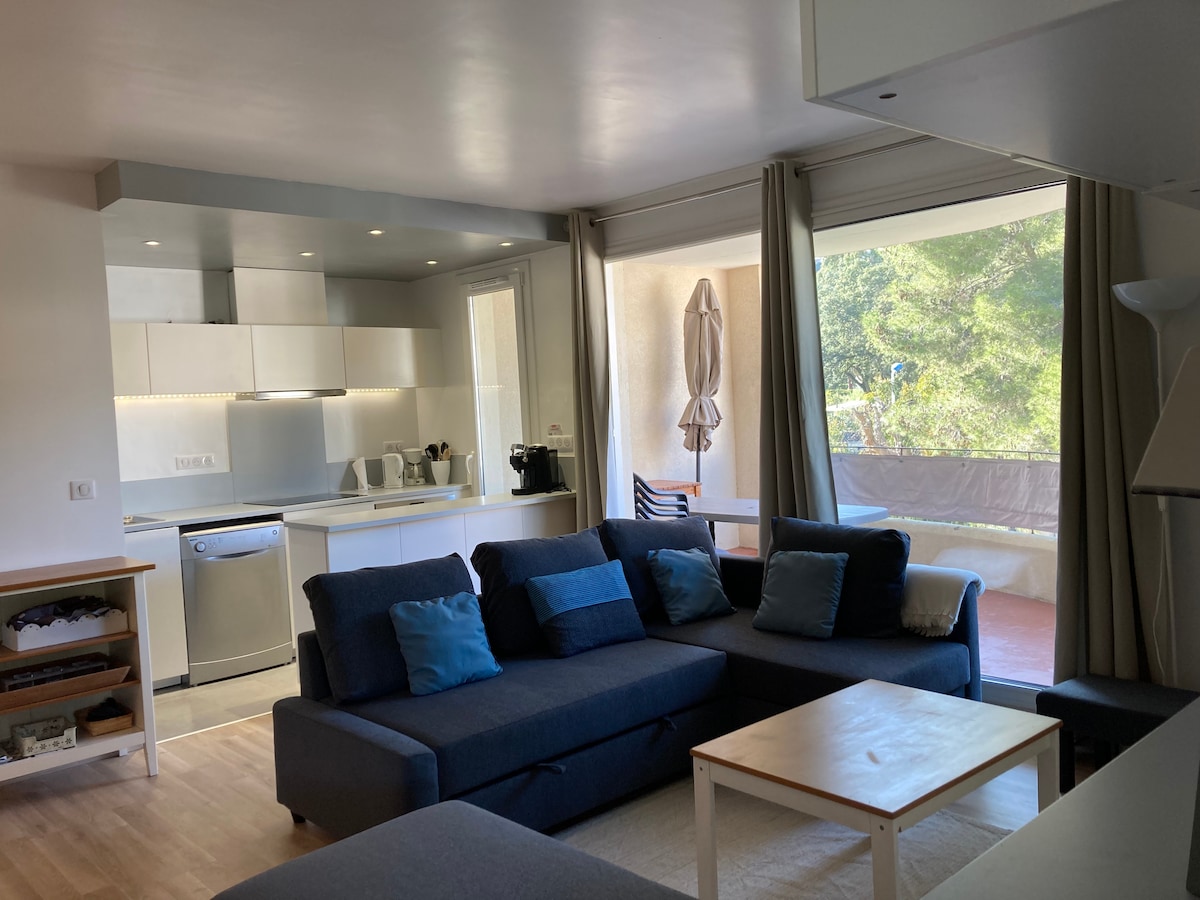 The living area features a spacious dark blue sectional sofa adorned with decorative cushions. A light-colored coffee table is positioned in front. The modern kitchen is visible, equipped with stainless steel appliances and minimalist cabinetry. Large glass doors provide access to the terrace, enhancing natural light.