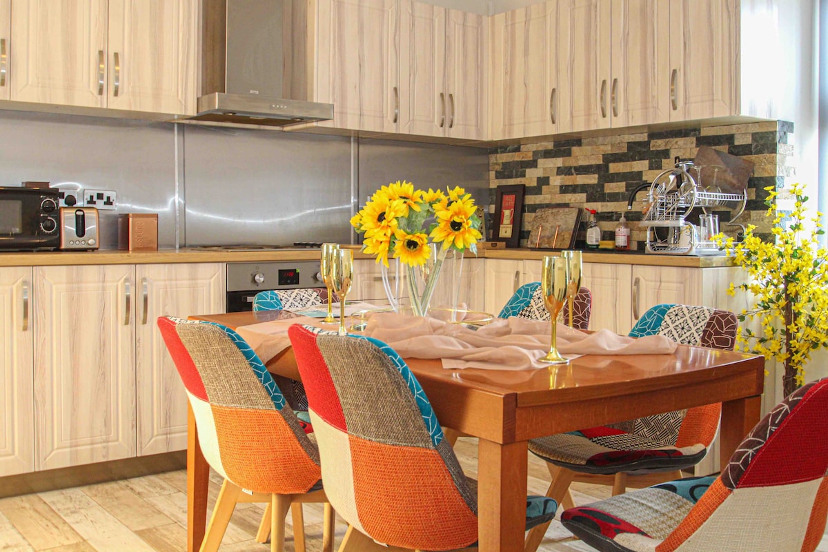 A modern kitchen is depicted, featuring a wooden dining table surrounded by colorful upholstered chairs. Sunflowers in a vase add a bright touch to the table setting, complemented by glasses and a neutral table cloth. Cabinetry and a subtle stone backsplash create a harmonious, stylish atmosphere.
