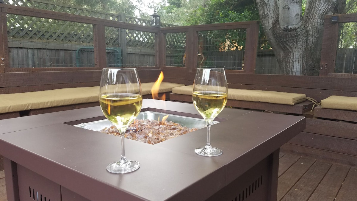 A firepit sits at the center of a wooden deck, surrounded by comfortable seating. Two glasses of white wine are placed on the edge of the firepit, with a small flame flickering nearby. A serene outdoor setting is framed by greenery and a wooden fence.
