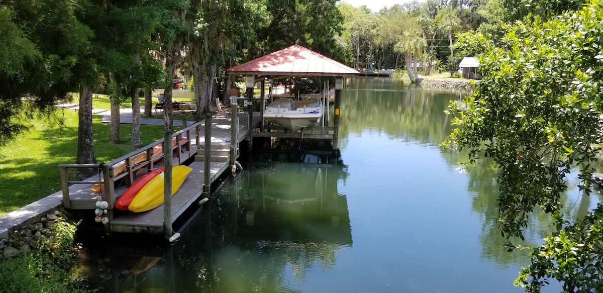 A serene canal is depicted with a wooden dock featuring a boat underneath a red-roofed shelter. Bright kayaks are positioned on the dock, surrounded by lush greenery and calm waters reflecting the trees. The tranquil atmosphere invites relaxation and outdoor exploration.