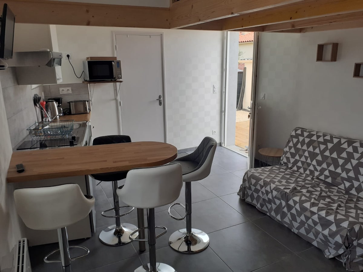 A functional kitchen area is depicted with a modern countertop and bar stools. A small dining table is visible alongside a cozy seating area, featuring a patterned sofa. Bright natural light enters from an open door, leading to an outdoor space.