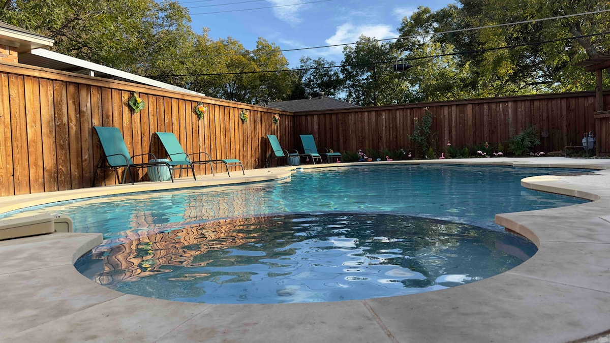 A tranquil swimming pool is set within a landscaped outdoor area, featuring a circular hot tub seamlessly integrated into the main pool. Multiple lounge chairs are positioned around the water, providing a space for relaxation under a clear sky. Natural greenery borders the area.