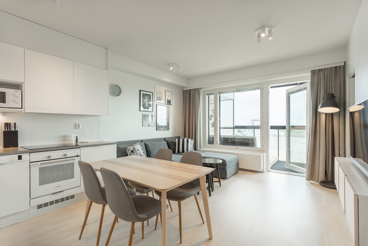 A modern living space features a fully equipped kitchen with sleek appliances. A dining table for four is set beside a spacious sofa and large windows leading to the balcony. Natural light fills the room, highlighting the neutral tones and minimalist décor.