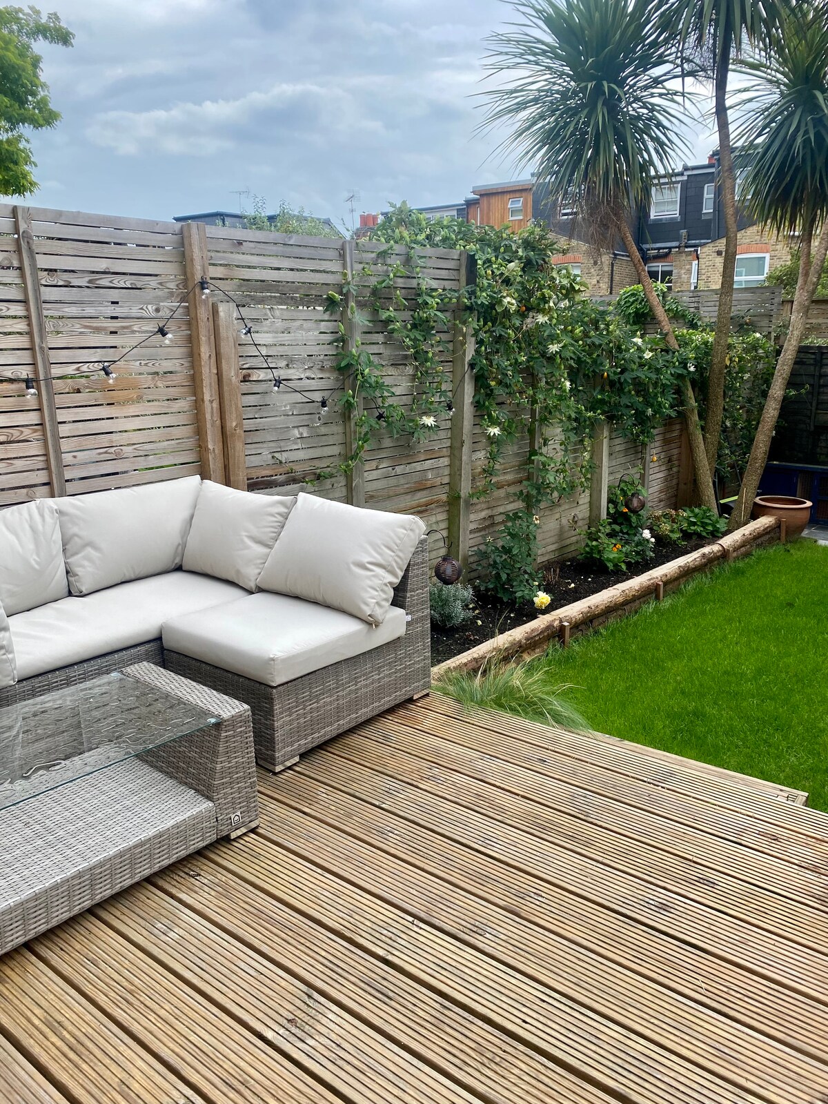 An outdoor terrace features a comfortable lounge area with soft, light-colored cushions and a glass-topped table. Lush greenery, including palm trees and climbing plants, adorns a wooden fence, while a neatly kept lawn provides a refreshing touch to the space.
