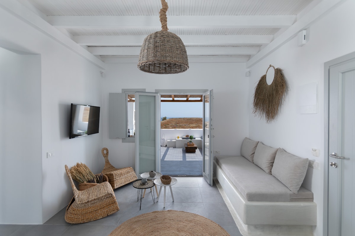 A bright living space is highlighted by large glass doors opening to a scenic view. A spacious sofa and woven chairs are arranged around a simple coffee table. Natural textures are added with a round jute rug and a hanging wicker light fixture.