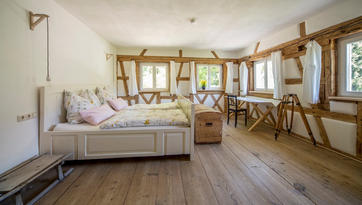 A spacious room features a light-colored bed adorned with floral linens and soft pillows. Natural light fills the area through multiple windows framed by wooden accents. A wooden table and two chairs are positioned nearby, complemented by a vintage trunk and a simple wooden bench.