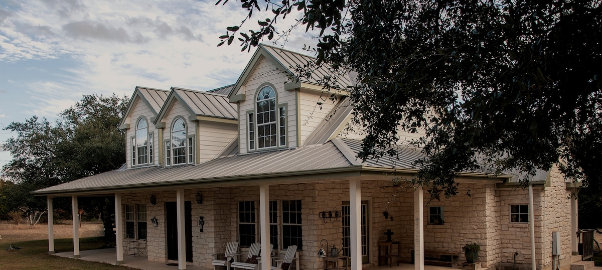 A charming two-story country home features a light exterior and a metal roof. Large windows provide ample natural light, while a covered porch with rocking chairs invites relaxation. Lush greenery surrounds the home, enhancing the serene landscape of the five-acre property.