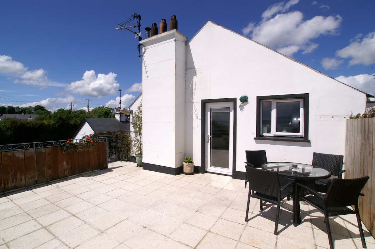 An enclosed patio area features a round table with four chairs, ideal for outdoor dining. Sunlight brightens the space, while a white wall and chimney complement the scene. Lush greenery is visible beyond the boundaries of the patio, contributing to a serene environment.