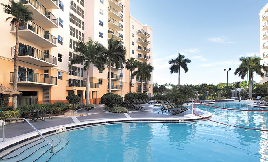 A large swimming pool surrounded by lush palm trees and greenery is visible. The pool area features several lounge chairs arranged around the water's edge. The backdrop includes a multi-story building with balconies, suggesting easy access to the outdoor amenities.