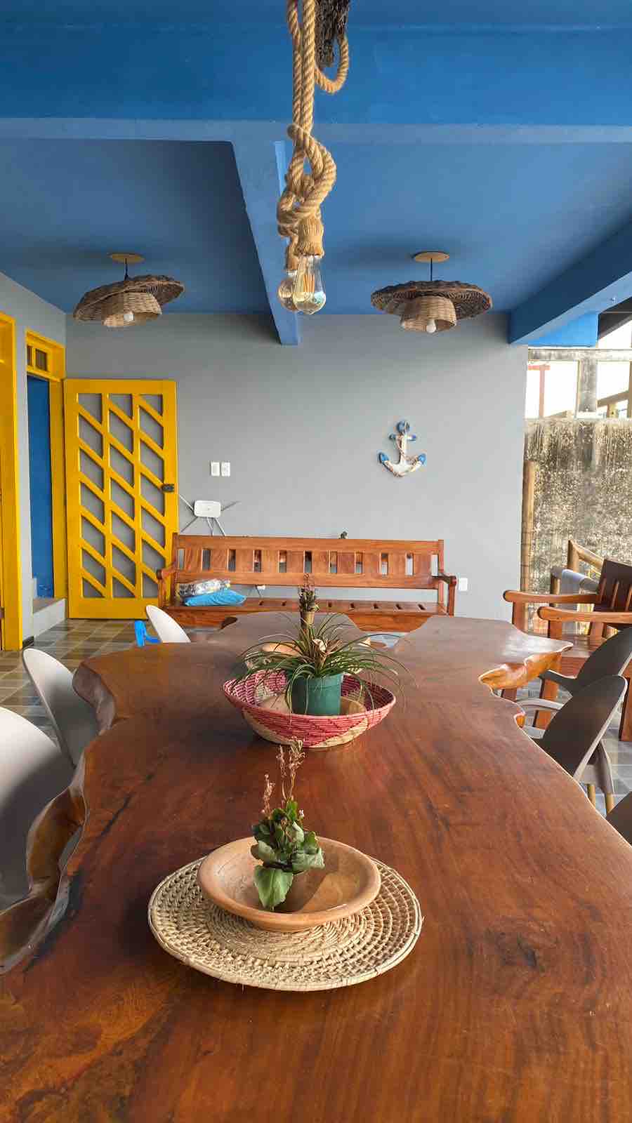 A long wooden dining table is centered in the image, adorned with decorative bowls and small plants. Surrounding chairs are visible, and colorful pendant lights hang from the blue ceiling. A wooden bench rests against the wall, and a nautical theme is suggested by decor items.
