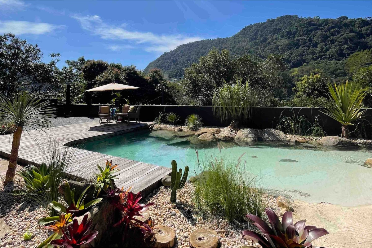 A natural swimming pool is surrounded by lush greenery and colorful plants. Wooden decking invites relaxation, and comfortable lounge chairs sit beneath a shade umbrella. Rocky formations enhance the serene landscape, while distant hills provide a scenic backdrop under a clear blue sky.