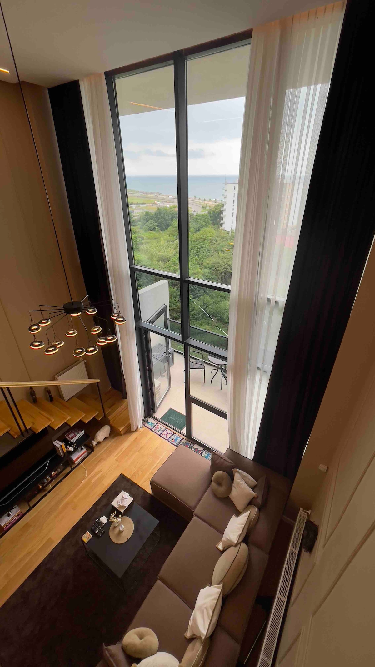The living area features high ceilings and large windows, allowing natural light to illuminate the space. A comfortable sofa is positioned in front of a low table, while decorative items add visual interest. A glimpse of greenery and distant views are visible through the glass.