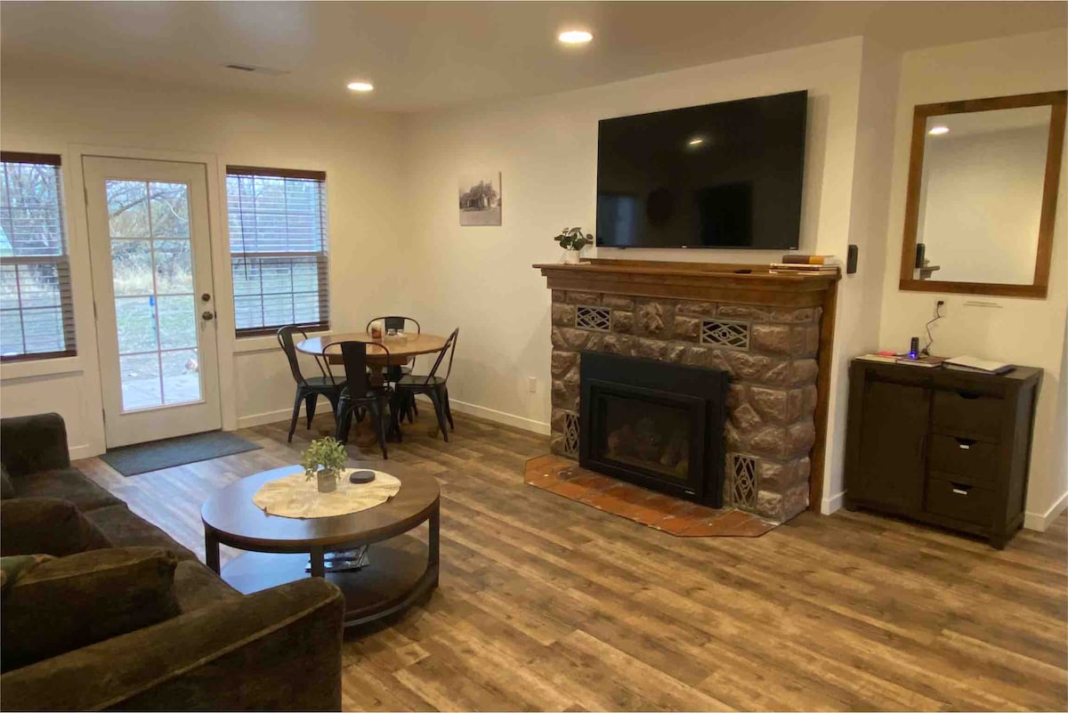 An open living area features a comfortable seating arrangement with a dark sofa and a round coffee table. A stone fireplace serves as a focal point under a mounted flat-screen TV. Dining space is provided with a small table and four chairs near large windows, allowing natural light in.
