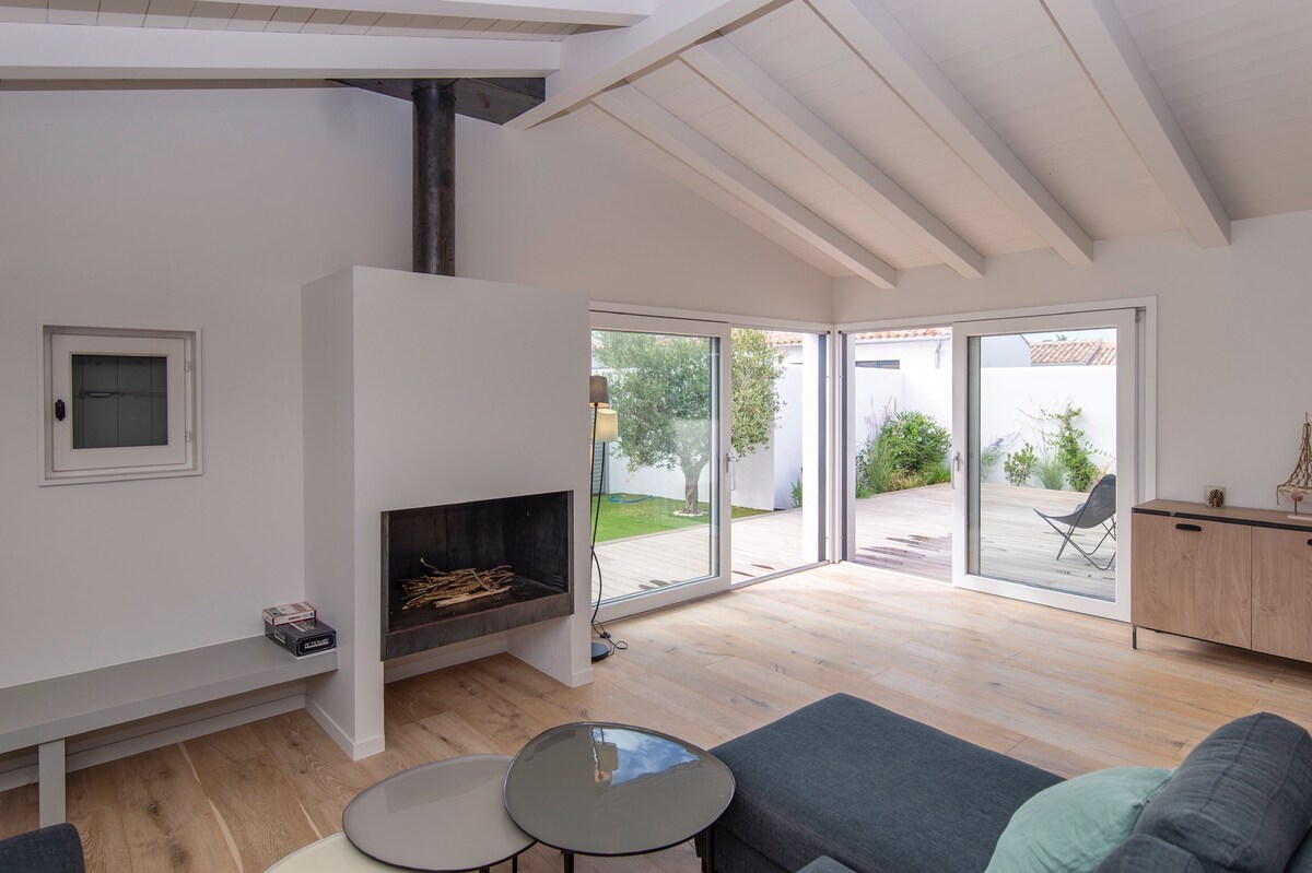 A modern living area is highlighted by a wood-burning fireplace and large glass doors that invite natural light. The sloped ceiling enhances the spacious feel, while wooden flooring adds warmth. A comfortable seating arrangement faces a sleek coffee table, creating an inviting space for gatherings.