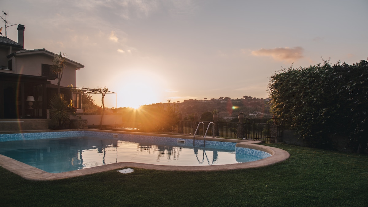 A clear swimming pool reflects the golden hues of the setting sun, surrounded by lush green grass and palm trees. The villa is positioned in the background, providing a serene view of the landscape beyond the fence.