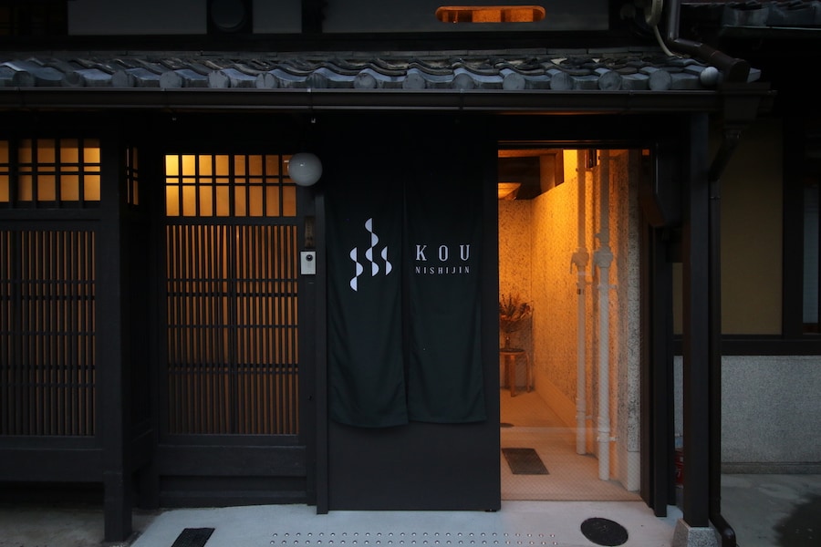 The entrance of KOU Nishijin features traditional wooden doors adorned with a welcoming sign. Soft lighting creates a serene ambiance, highlighting the architectural details of the exterior. The space is framed by textured walls and a tiled pathway leading inside.