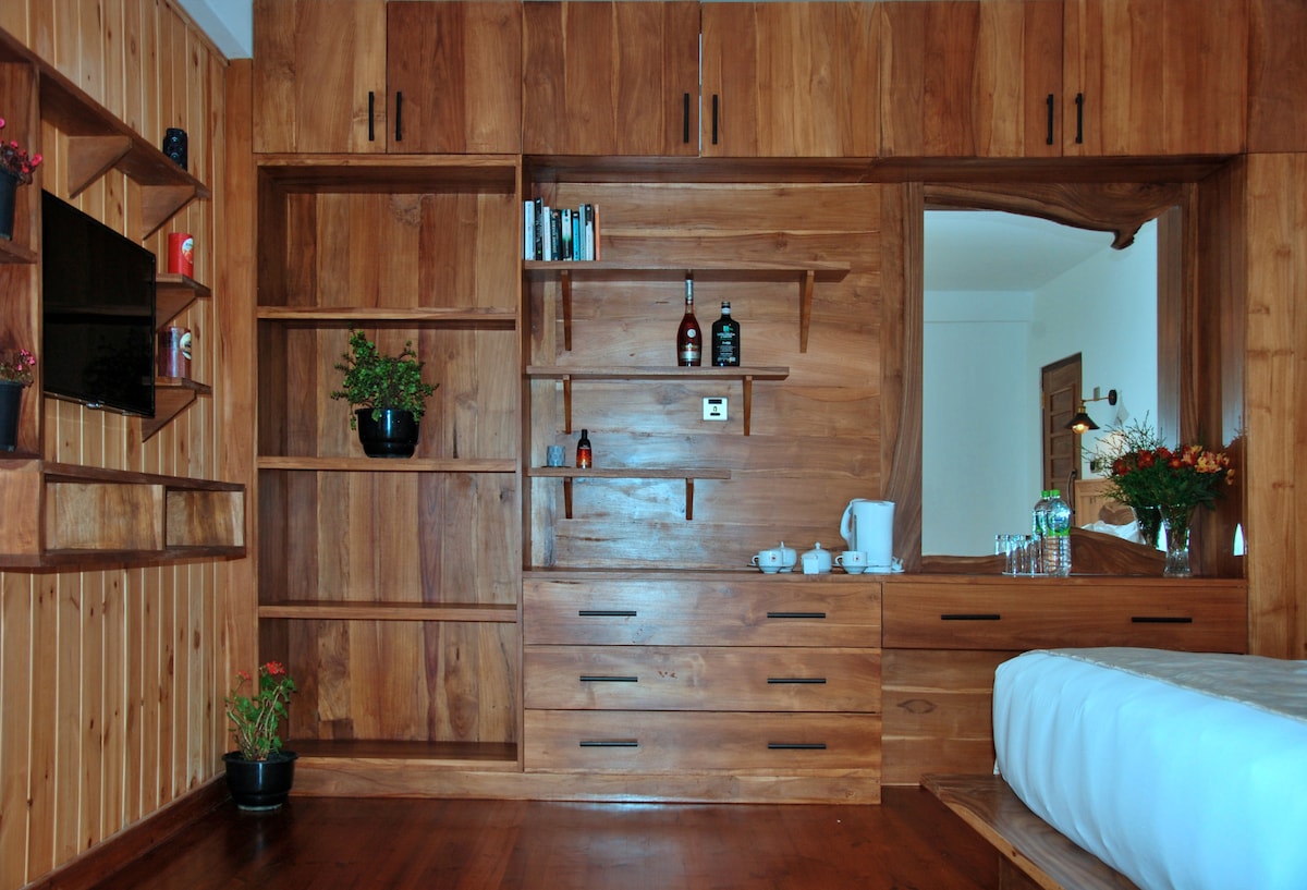 A warm wooden wall unit displays shelves and drawers, creating ample storage space. Below, a sleek countertop holds a tea set and glasses. A small plant and flowers add touches of greenery, while a flat-screen TV is mounted for convenience.