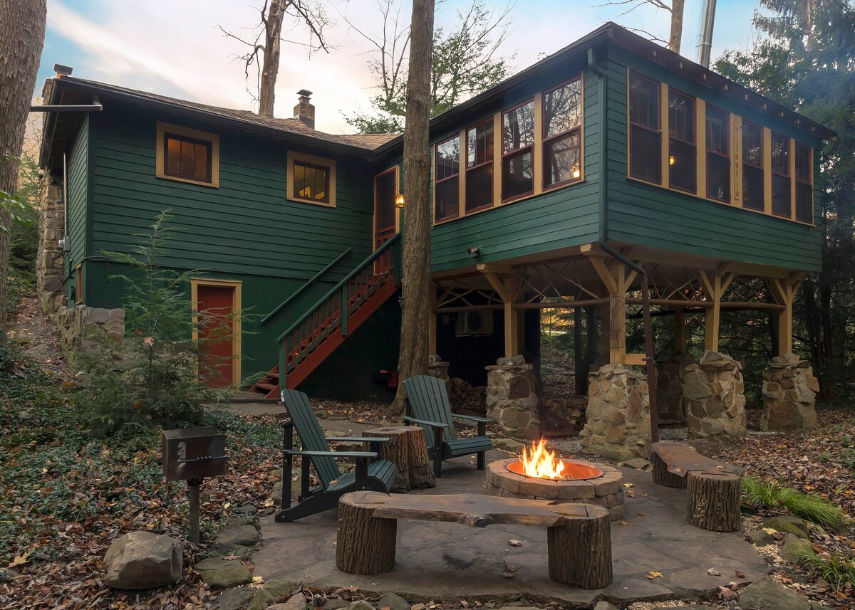 A welcoming outdoor fire pit area is surrounded by rustic stone seating. Nearby, two adirondack chairs invite relaxation in a serene, wooded setting. The green cottage features large windows on the upper level, providing views of the natural surroundings.