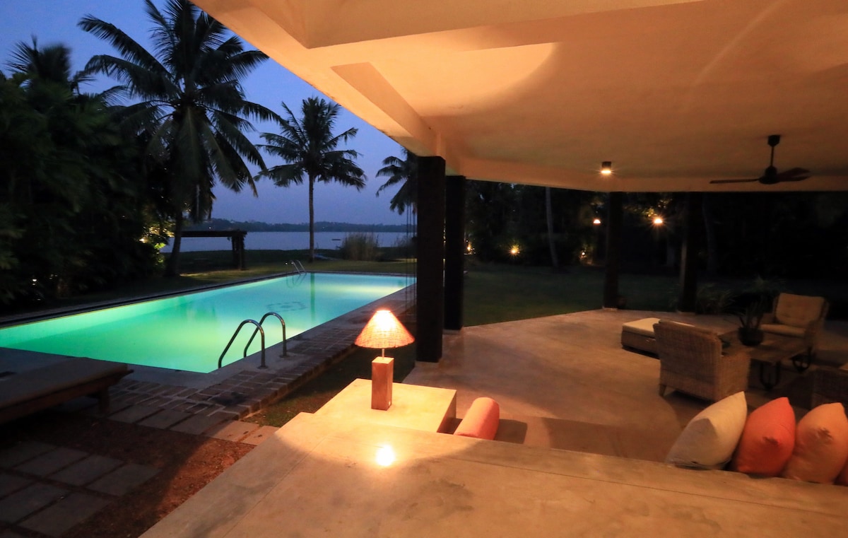 An outdoor seating area is showcased beside a shimmering pool, with tropical palm trees in the background. Soft lighting illuminates the space, creating a calming ambiance. The pool reflects the twilight sky, while comfortable lounge chairs invite relaxation.