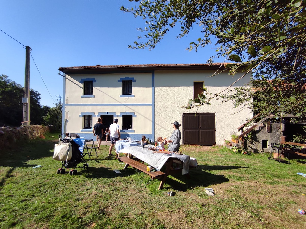 The exterior of the house features a light-colored facade with blue window frames and a large brown door. A picnic table occupies the yard, surrounded by a grassy area. Guests can be seen enjoying the outdoor space, with trees providing partial shade.