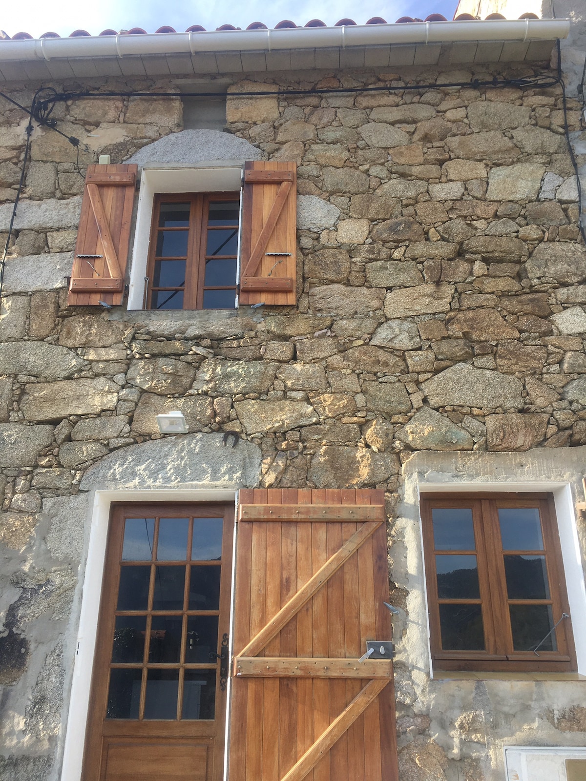 The exterior of the stone cottage showcases traditional architecture with weathered stone walls and wooden shutters. An entrance door with glass panes complements a rustic wooden door on the lower level. Natural light is captured through the upper window, revealing a harmonious blend with the surrounding landscape.