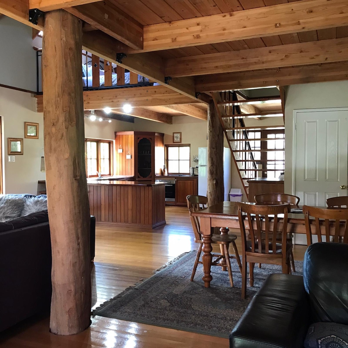 The spacious open-plan living area is highlighted by exposed wooden beams and polished timber flooring. A comfortable seating arrangement is positioned near a dining table, while a staircase leads to an upper mezzanine. The kitchen, featuring wooden cabinetry, is visible in the background.