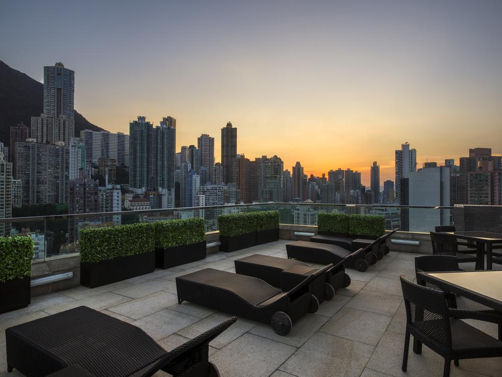 A rooftop terrace is equipped with several lounge chairs facing a panoramic view of the Hong Kong skyline at sunset. The scene captures skyscrapers silhouetted against the fading light, with planters providing greenery along the edge of the terrace.