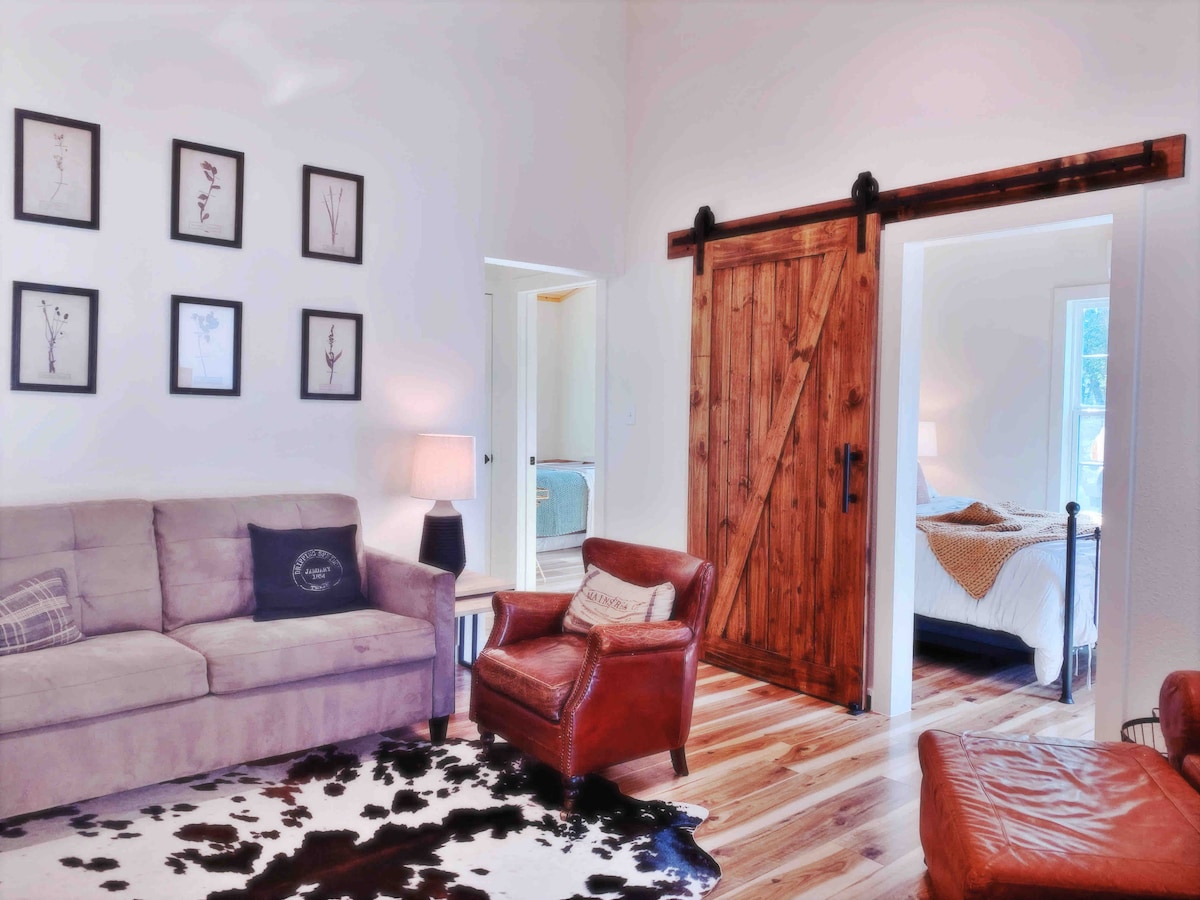 A cozy living space features a light-colored sofa and a rustic armchair. Artwork is displayed on the walls, and a wooden barn door adds character. Natural light fills the room, creating a warm and inviting ambiance for relaxation.
