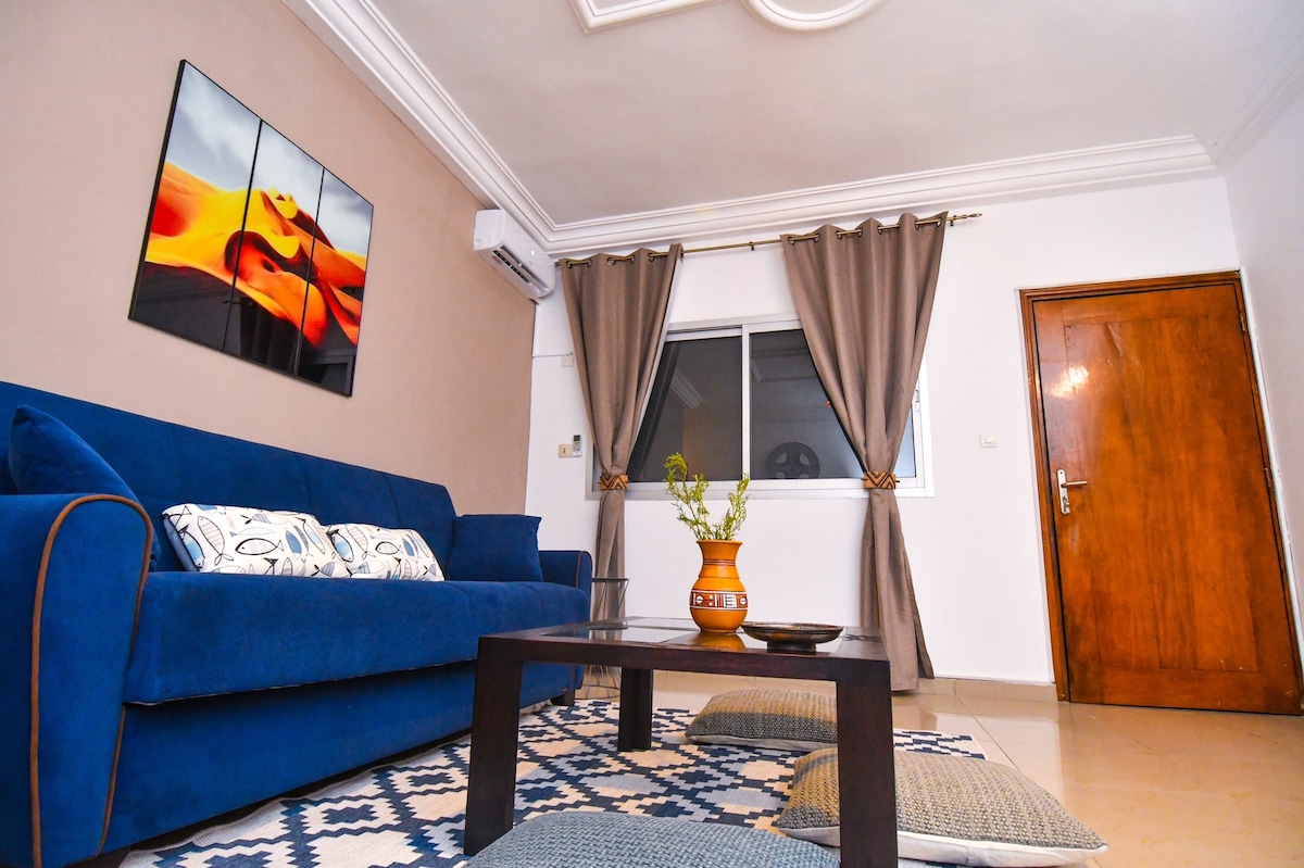 A cozy living area is highlighted by a blue sofa adorned with decorative pillows. A wooden coffee table sits in the center, accompanied by floor cushions. A large piece of abstract art hangs on the wall, and natural light enters through the nearby window, framed by soft curtains.