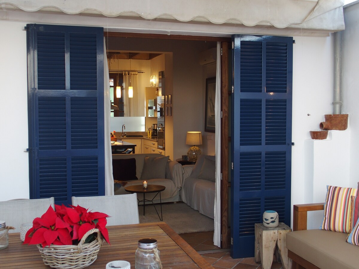 Blue shutters frame an inviting entrance to a cozy living space. Soft furnishings are visible in the background, while a dining table set with potted plants and candles occupies the outdoor area, creating a welcoming atmosphere for gatherings.